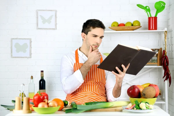 Homem com livro de receitas em mãos à mesa com diferentes produtos e utensílio na cozinha no fundo da parede branca — Fotografia de Stock