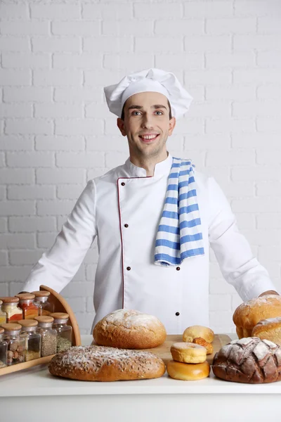 Baker in kitchen at table with freshly loaves of bread on white brick wall background — Stock Photo, Image