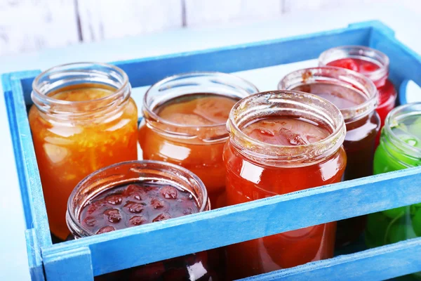 Homemade jars of fruits jam in crate on table and color wooden planks background — Stock Photo, Image