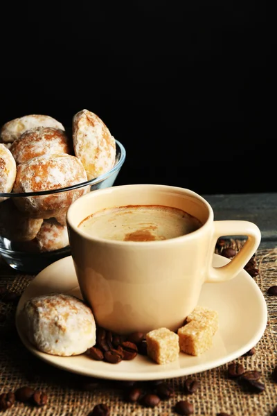 Cup of coffee and tasty cookies on sackcloth napkin, on wooden table, on dark background — Stock Photo, Image