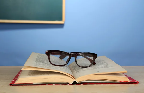 Livre ouvert avec des lunettes sur un bureau en bois, sur un mur coloré et fond de tableau noir — Photo