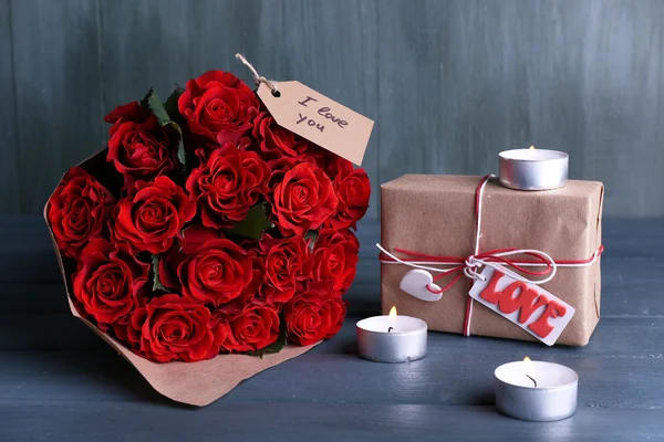 Bouquet of red roses wrapped in paper with present box and candles on wooden background — Stock Photo, Image