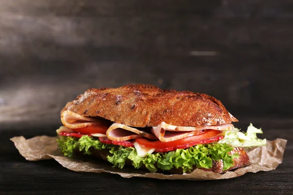 Fresh and tasty sandwich with ham and vegetables on paper on wooden background — Stock Photo, Image