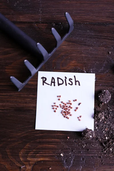 Radish seeds on piece of paper with ground and rake on wooden background — Stock Photo, Image