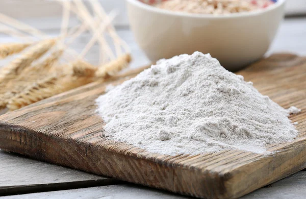 Heap of flour on cutting board with ears and grains in bowl on wooden table — Stock Photo, Image