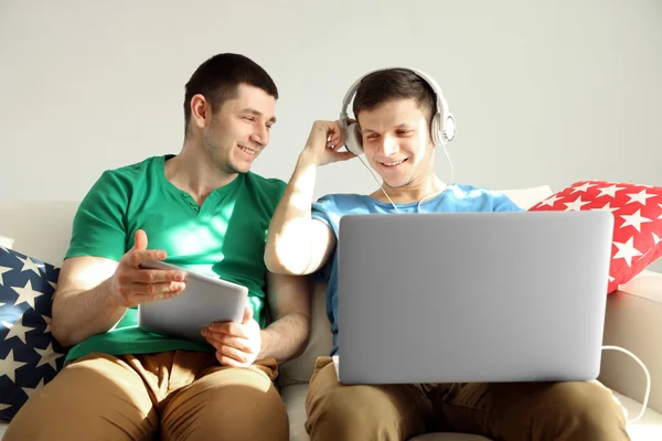 Two handsome young men sitting on sofa and using gadgets — Stock Photo, Image