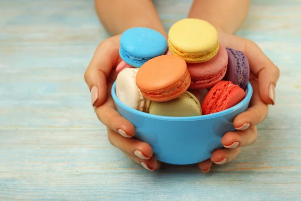 Macarons colorés savoureux dans un bol et mains féminines sur fond de bois de couleur — Photo