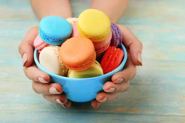 Macarons colorés savoureux dans un bol et mains féminines sur fond de bois de couleur — Photo