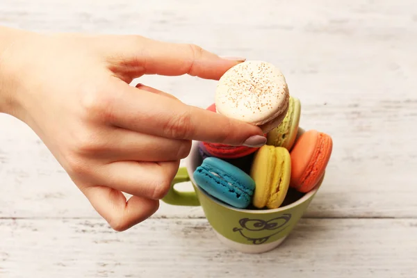 Main féminine prenant des macarons colorés savoureux de tasse sur fond en bois de couleur — Photo