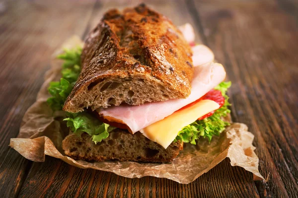 Fresh and tasty sandwich with ham and vegetables on paper on wooden background — Stock Photo, Image