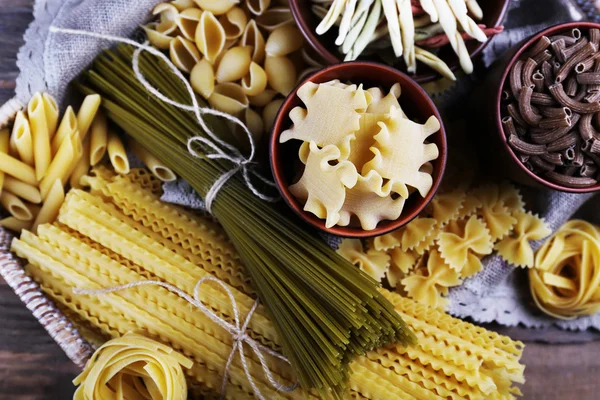 Verschillende soorten pasta in rieten mand met stof op rustieke houten tafel achtergrond — Stockfoto