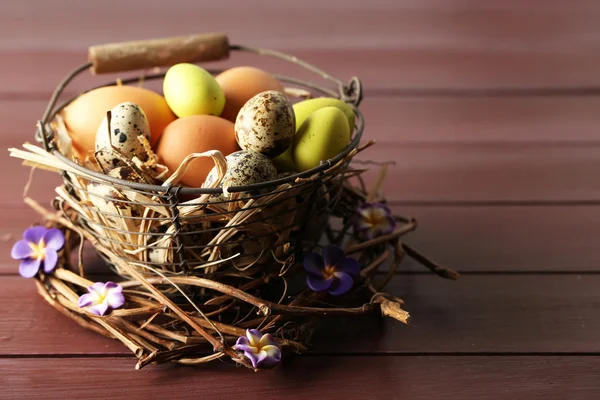 Птичьи яйца в плетеной корзине с декоративными цветами на фоне цвета дерева — стоковое фото