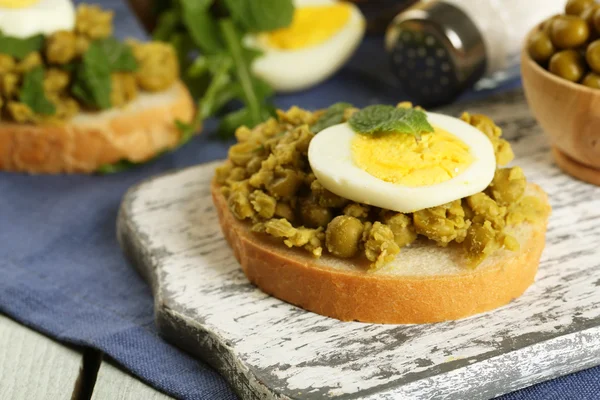 Sandwiches with green peas paste and boiled egg on cutting board with napkin on color wooden planks background — Stock Photo, Image