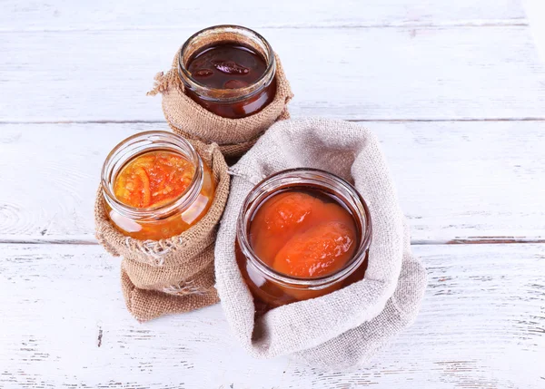 Homemade jars of fruits jam in burlap pouches on color wooden planks background — Stock Photo, Image