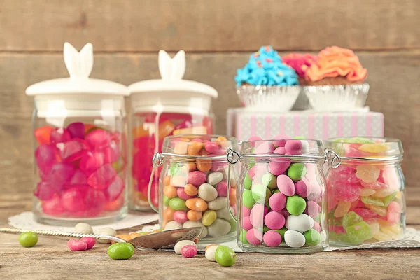 Mehrfarbige Bonbons in Glasgläsern und Cupcakes auf Holzgrund — Stockfoto