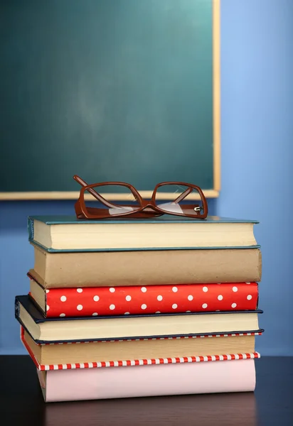Empilement de livres avec des lunettes sur un bureau en bois, sur un mur coloré et fond de tableau noir — Photo