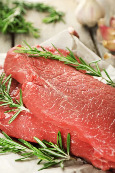 Raw beef steak with rosemary and garlic on paper on wooden background — Stock Photo, Image