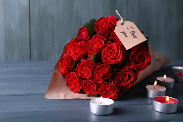 Boeket van rode rozen met tag gewikkeld in papier en kaarsen op houten achtergrond — Stockfoto