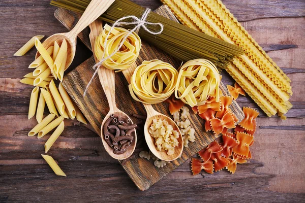 Verschillende soorten pasta op snijplank en houten tafel achtergrond — Stockfoto