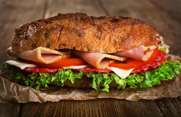 Sandwich fresco y sabroso con jamón y verduras sobre papel sobre fondo de madera — Foto de Stock