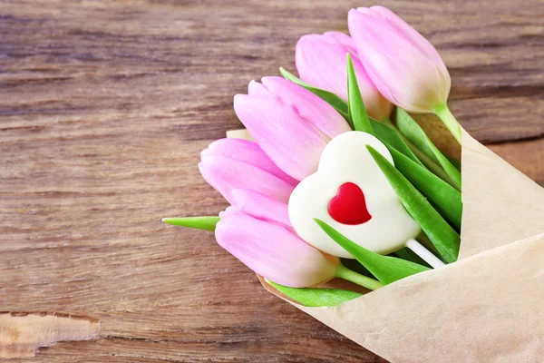 Mooie roze tulpen in papier met zoete hart op houten achtergrond — Stockfoto