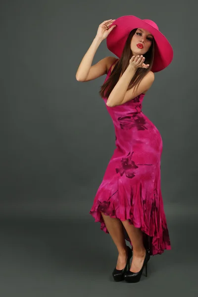 Красива молода жінка в довгій рожевій сукні і капелюсі на темно-сірому фоні — стокове фото