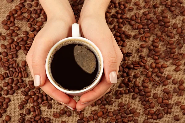 Mains féminines tenant la tasse avec des grains de café sur toile de jute, vue de dessus — Photo