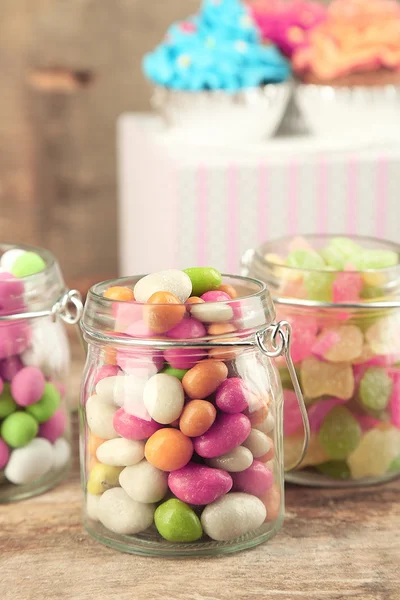 Многоцветные конфеты в стеклянных банках и кексах на деревянном фоне — стоковое фото