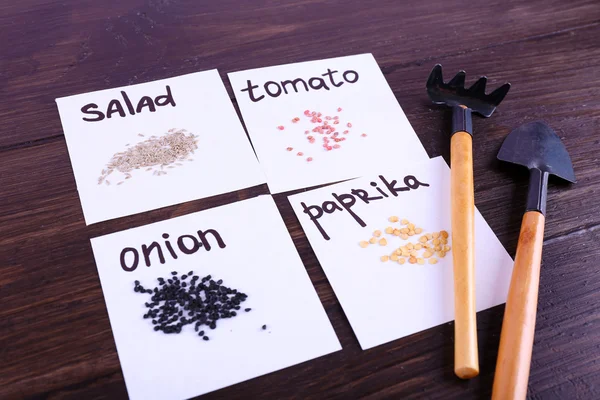 Different seeds on pieces of paper with gardening tools on wooden background — Stock Photo, Image