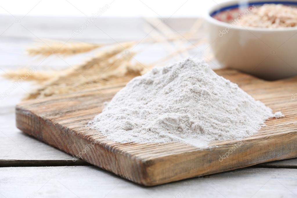 Heap of flour on cutting board with ears and grains in bowl on wooden table