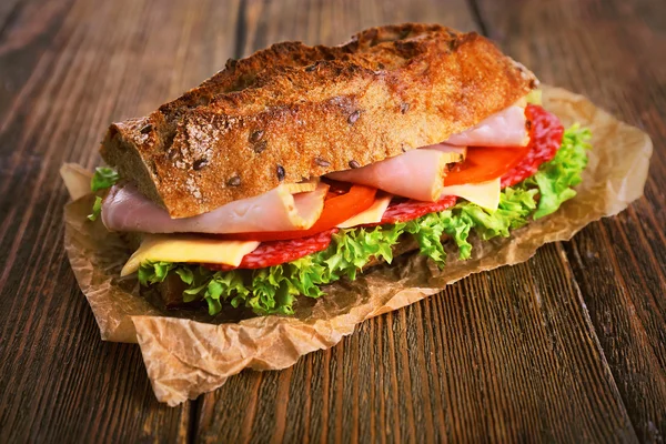 Fresh and tasty sandwich with ham and vegetables on paper on wooden background — Stock Photo, Image