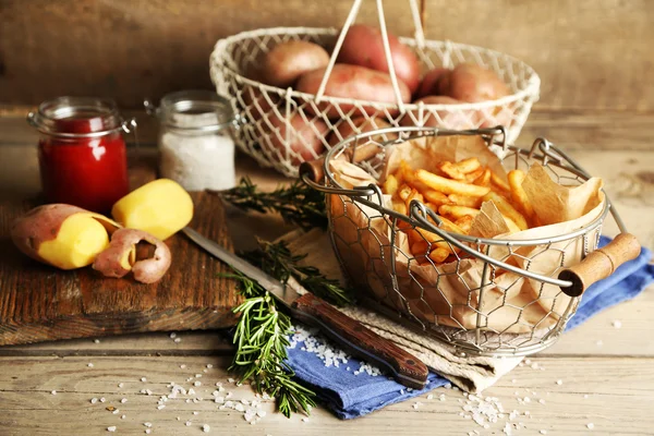 Tasty french fries and fresh potatoes in metal baskets on wooden table background — Stock Photo, Image