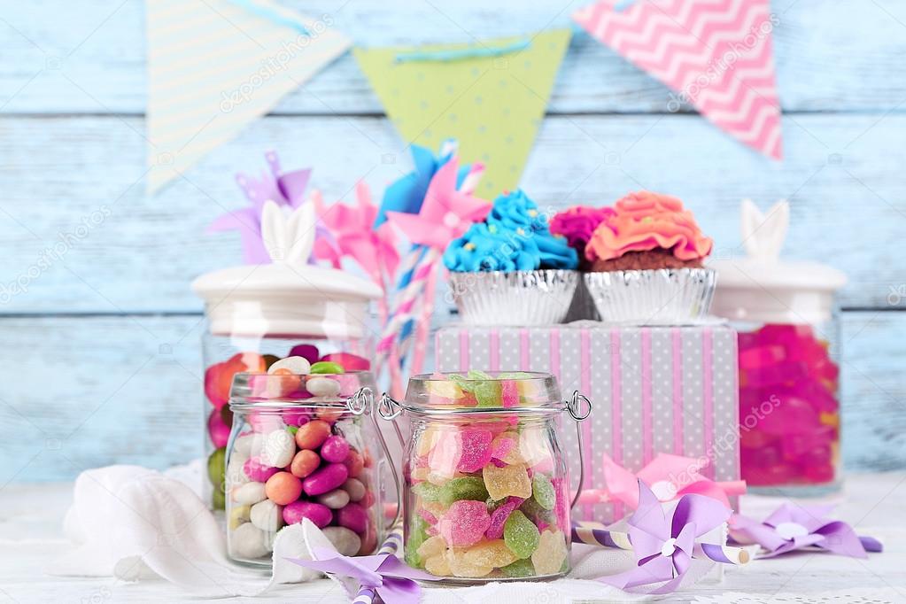 Multicolor candies in glass jars and cupcakes on color wooden background