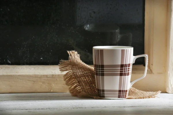 Cup of hot drink with napkin on windowsill on rain background — Stock Photo, Image