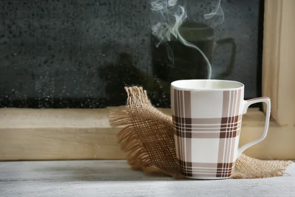 Cup of hot drink with napkin on windowsill on rain background — Stock Photo, Image