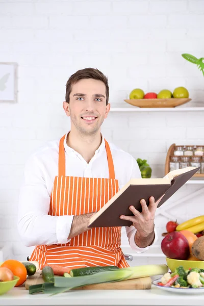 Человек с книгой рецептов в руках за столом с различными продуктами и посудой на кухне на белом фоне стены — стоковое фото