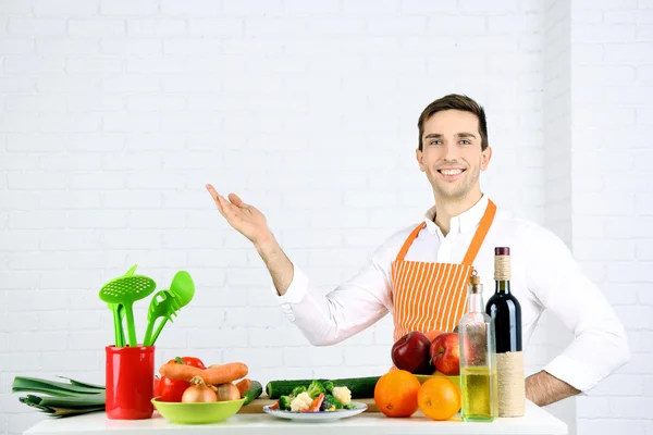 Mann am Tisch mit verschiedenen Produkten und Utensilien in der Küche auf weißem Wandhintergrund — Stockfoto