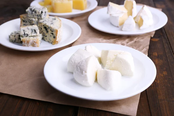 Plates of various types of cheese on sheet of paper on wooden table background — Stock Photo, Image