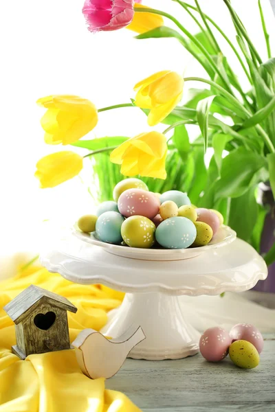 Huevos de Pascua en jarrón y tulipanes en mesa sobre fondo brillante —  Fotos de Stock