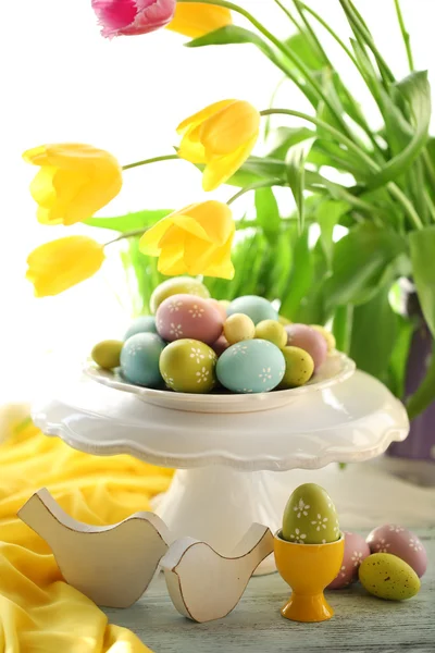 Huevos de Pascua en jarrón y tulipanes en mesa sobre fondo brillante —  Fotos de Stock