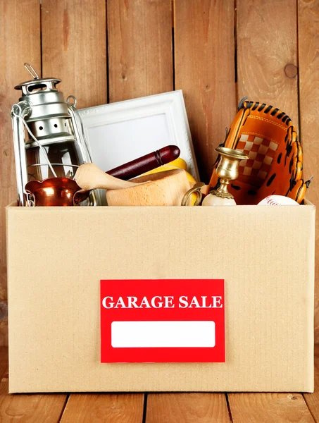 Box of unwanted stuff ready for a garage sale on wooden background — Stock Photo, Image