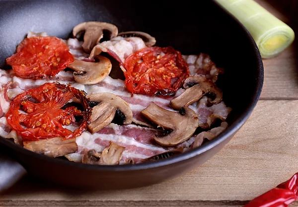 Strips of bacon with sliced mushrooms and tomato in pan on wooden planks background — Stock Photo, Image