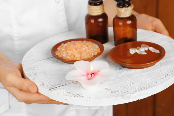 Beauty therapist holding tray of spa treatments, close-up, on wooden wall background — Stock Photo, Image