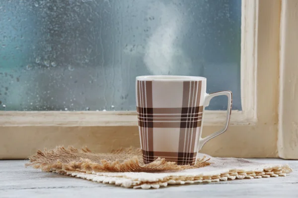 Cup of hot drink with napkin on windowsill on rain background — Stock Photo, Image