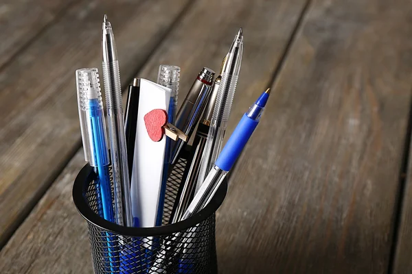 Verschillende pennen in metalen houder met aantekeningen op papier op houten planken achtergrond — Stockfoto