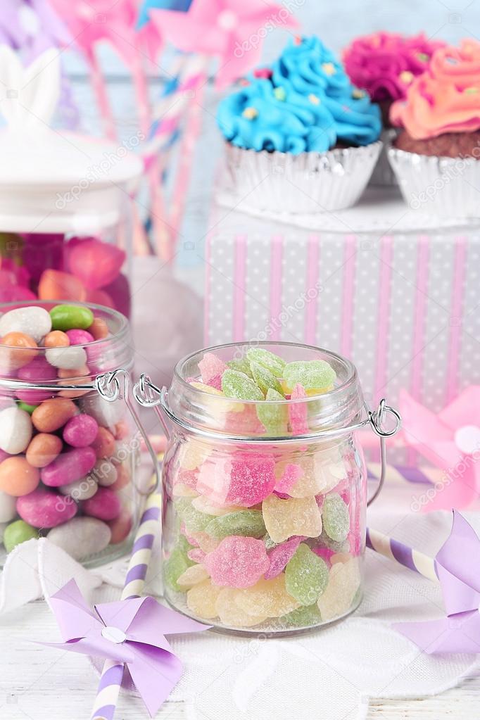 Multicolor candies in glass jars and cupcakes on color wooden background