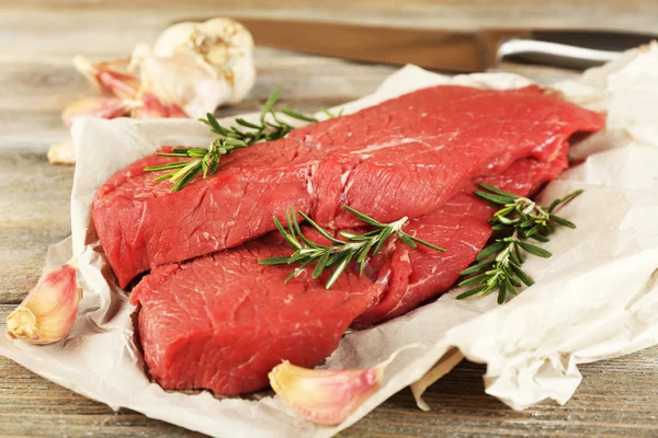 Raw beef steak with rosemary and garlic on paper on wooden background — Stock Photo, Image