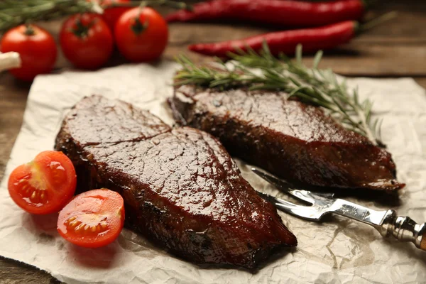 Složení s chutné pečené maso na papírový list, rajčata a rozmarýn větvičky na dřevěné pozadí — Stock fotografie
