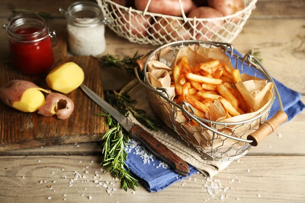 Tasty french fries and fresh potatoes in metal baskets on wooden table background — Stock Photo, Image