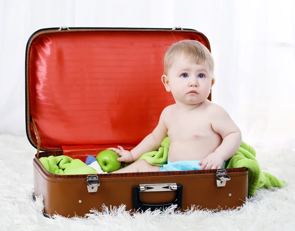 Lindo bebé niño sentado en una maleta grande en la alfombra, sobre fondo claro — Foto de Stock